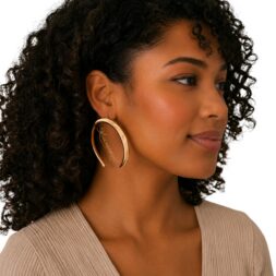 Woman wearing oversized gold tone open hoop earrings with ALOUMBABELLE® logo overlay inside the earrings on neutral background